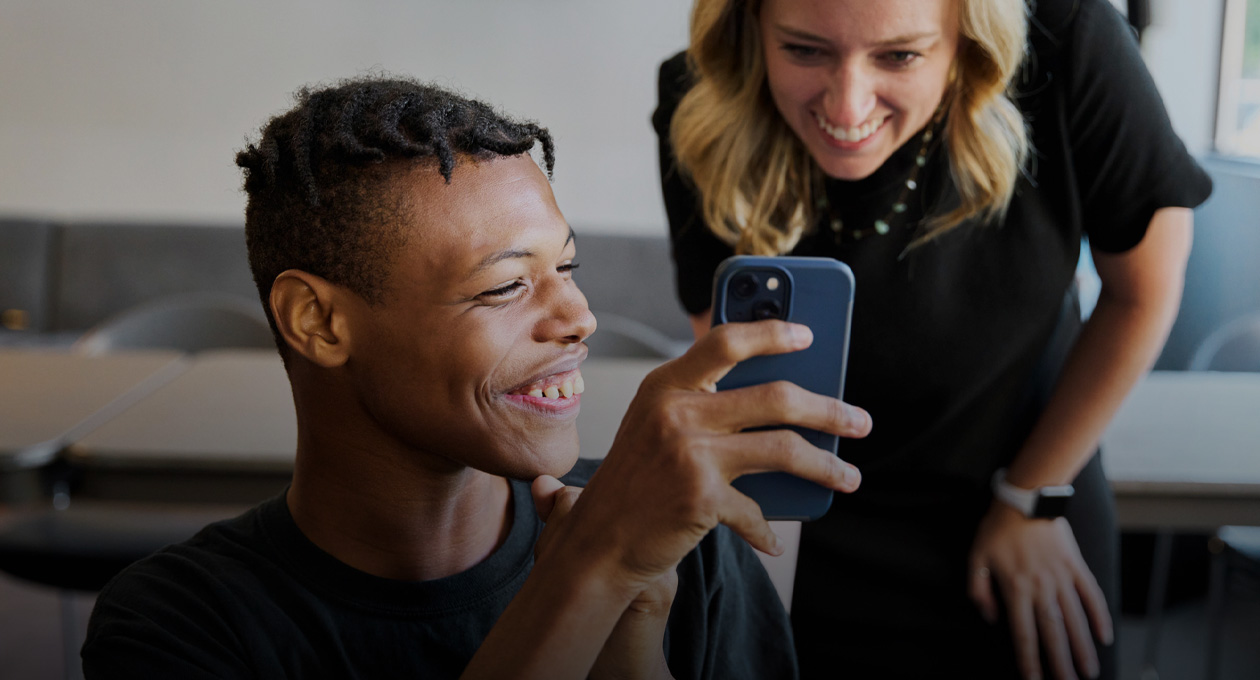 視覚に障がいのある若い男性の写真。左後ろに立っている笑顔の女性にiPhoneの画面を見せて微笑んでいる