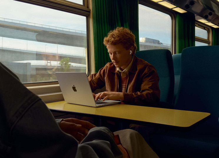 Una persona en el asiento de un tren junto a la ventana usa unos AirPods y trabaja en una MacBook.