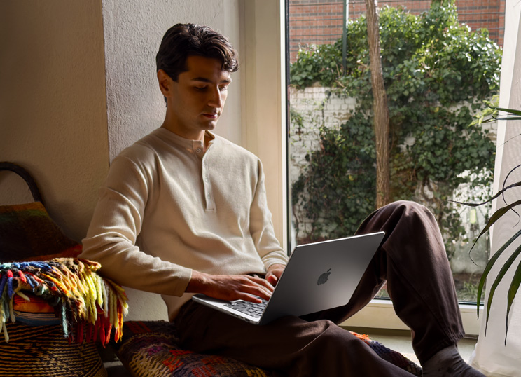 Una persona sentada en el suelo junto a una ventana está usando una MacBook.