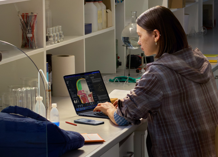 Una persona sentada en un laboratorio usa una MacBook; la pantalla muestra varias apps.