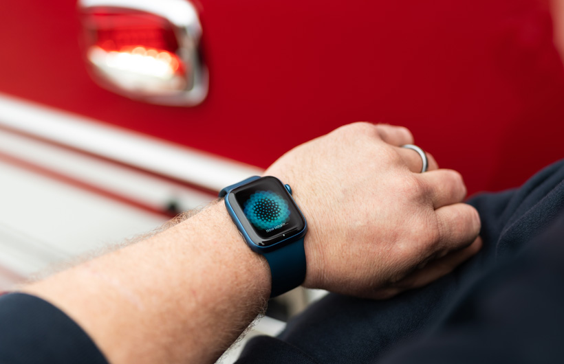 Một cánh tay đeo Apple Watch hiển thị thông báo: Giờ hãy hít vào, với một chiếc xe cứu hỏa ở gần bên