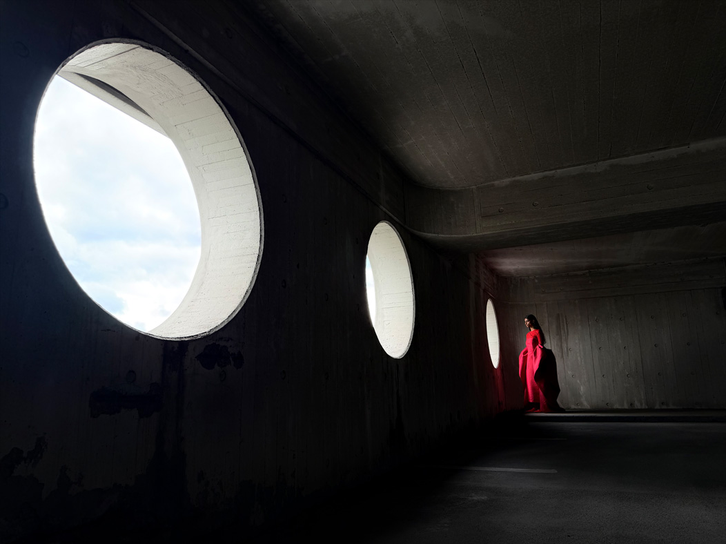 Una mujer con un largo vestido rojo de pie en medio de una habitación vacía mientras la fotografían. La distancia focal de la imagen se va alternando en la app Fotos para mostrar las capacidades del sistema de cámaras Pro: 24 mm